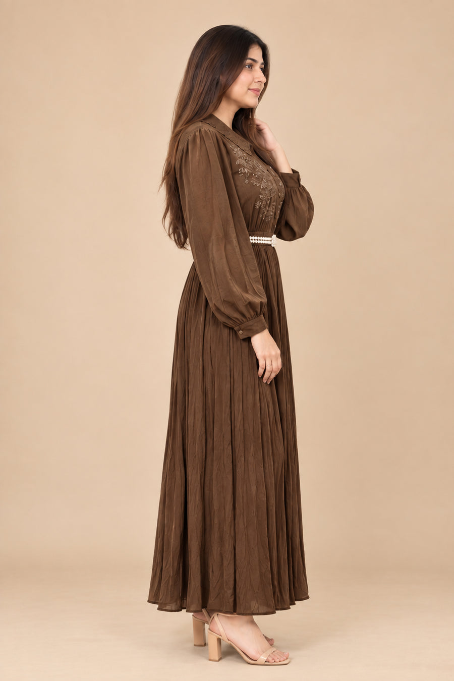 A woman modeling a long-sleeved brown maxi dress with a pleated skirt and lace bodice, standing against a solid beige studio background.